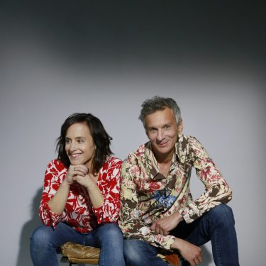 Laura und Tobias Goldfarb tragen beide bunte Hemden und sitzen nebeneinander auf einer Bank in einem leeren Raum